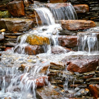 Water cascades over layered, reddish-brown rocks in a small, multi-tiered waterfall surrounded by smooth stones.
