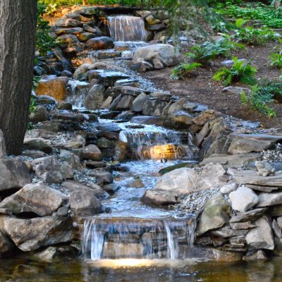 A small, tiered waterfall flows over rocks between two trees in a landscaped garden with accent lighting.