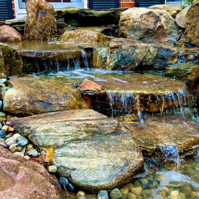 Water flows over tiered rocks in a landscaped garden stream surrounded by stones and greenery.