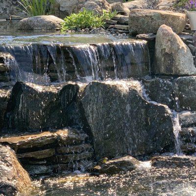Small waterfall cascades over rocks into a pond with a white flower pot and purple flowers on the right.