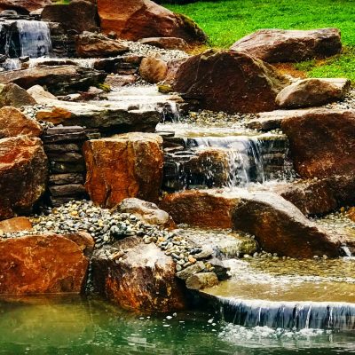 Small, multi-level waterfall flows over large rocks and pebbles into a pond, surrounded by grass and stones.