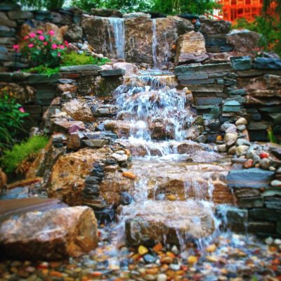 Small artificial waterfall flows over stacked rocks, surrounded by green plants and pink flowers in a landscaped garden.