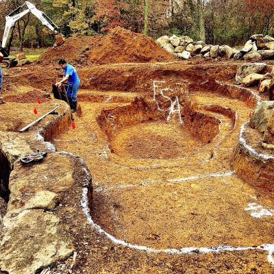 Two workers excavate a multi-level site outlined with white chalk, surrounded by rocks and construction equipment.